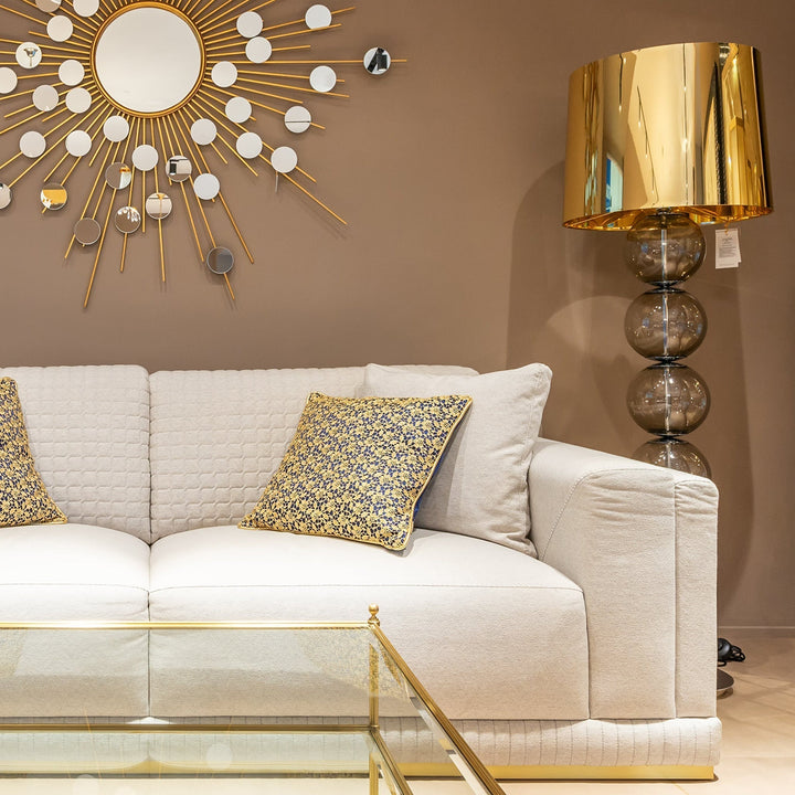 White sofa with a gold-patterned pillow under a sunburst mirror, beside a gold floor lamp