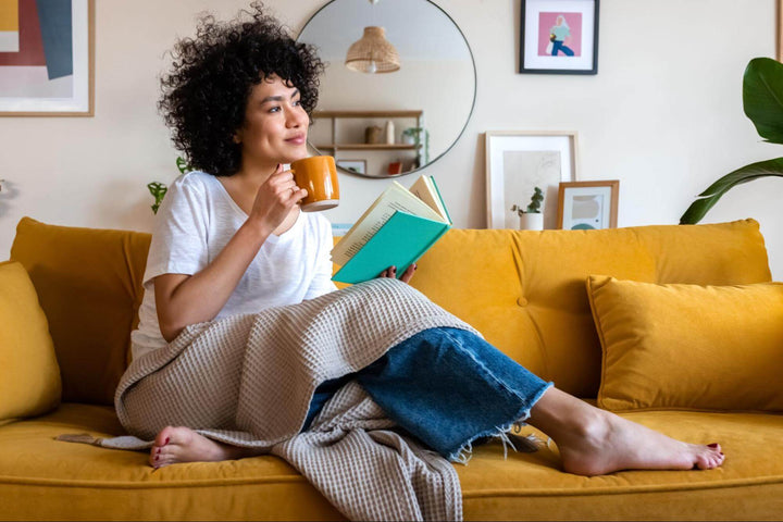 Woman with a mug and a book relaxing under a throw on a mustard yellow couch