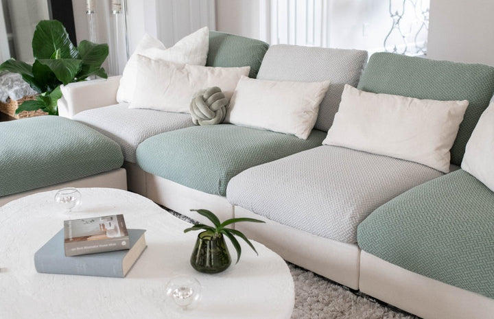 Sectional sofa with alternating sage green and light grey stretch cushion covers and a round white coffee table
