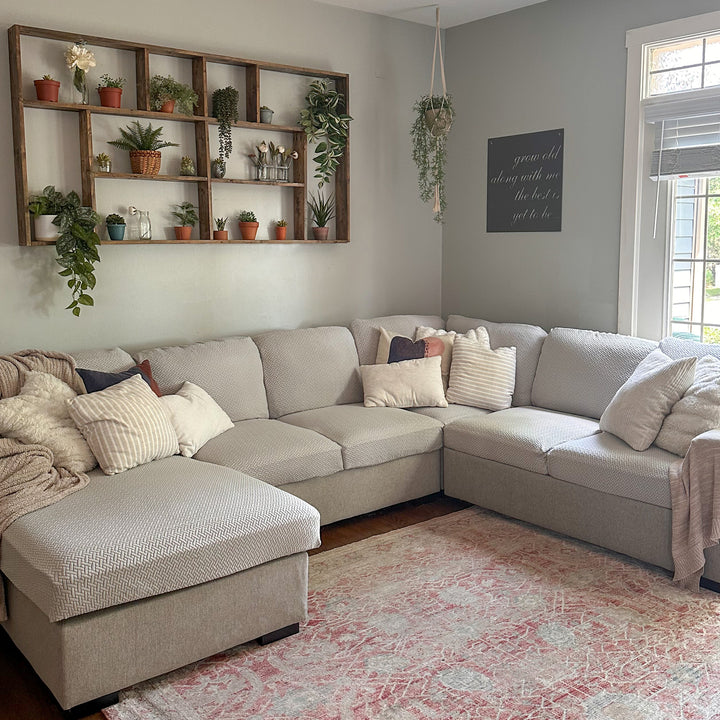 Sectional sofa with light grey textured stretch covers in a living room with a plant shelf and a pink rug
