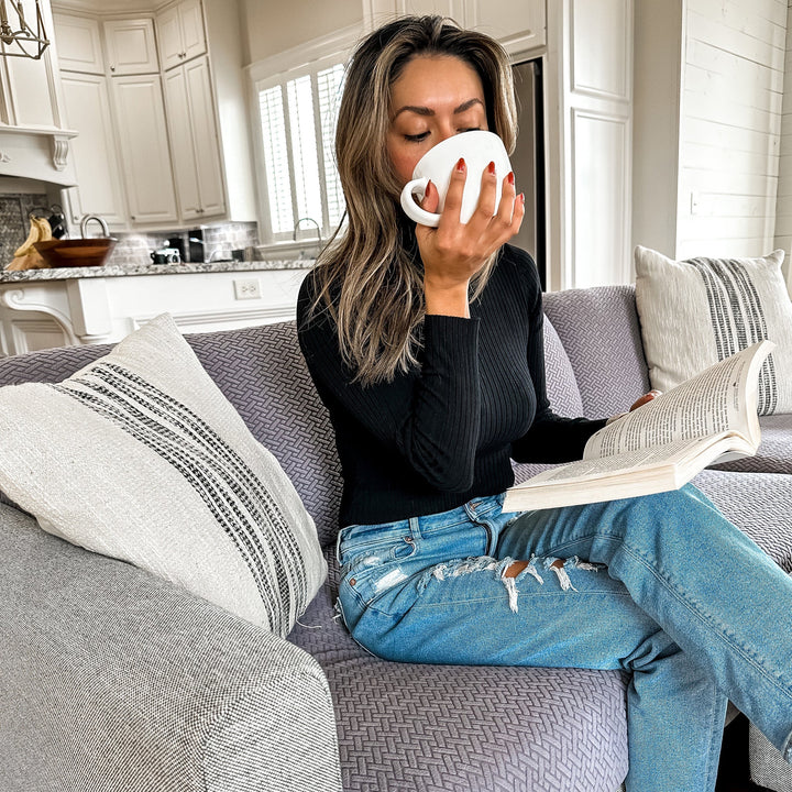 Woman with a mug and a book on a grey sectional with textured stretch covers and striped pillows