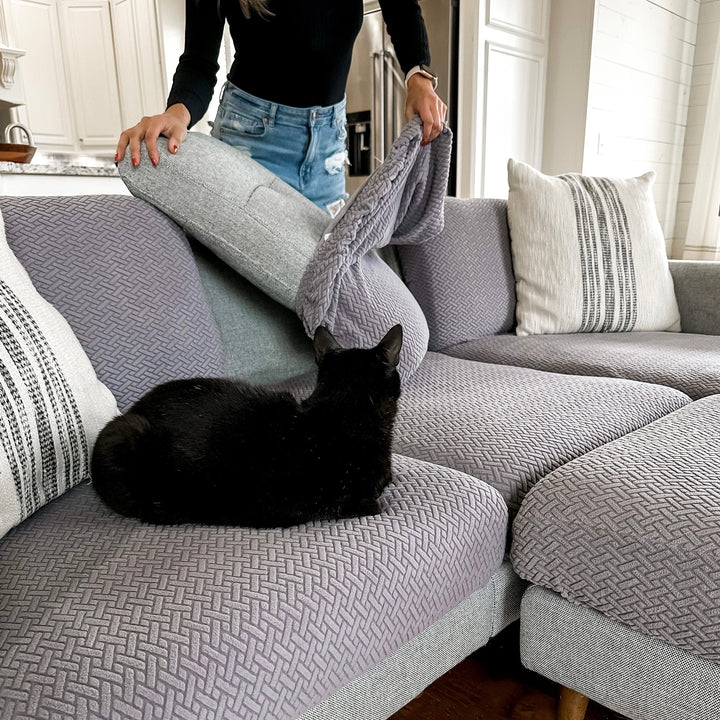 Woman fitting a grey cushion cover on a sectional as a black cat sits on a textured grey seat cover