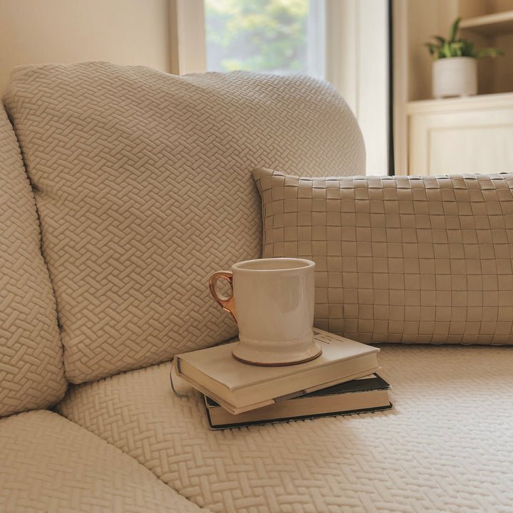 Beige textured stretch cover on a sofa with a woven lumbar pillow and a mug on a stack of books