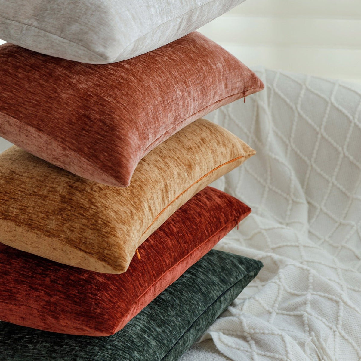 Stack of Woody pillow covers in grey, dusty rose, mustard, rust and dark green beside a white knit throw
