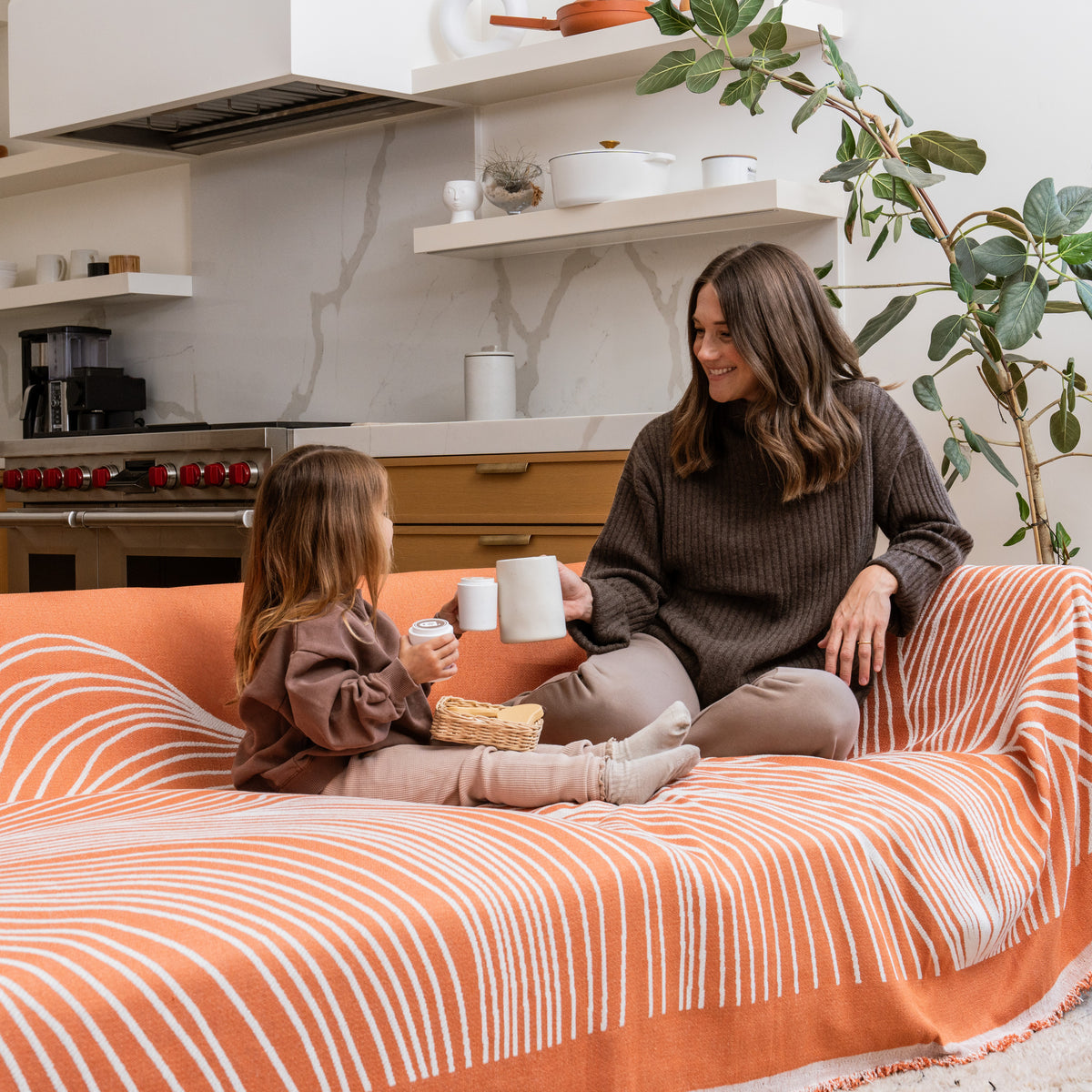 Woman and girl hold cups on a sofa draped in an orange patterned blanket